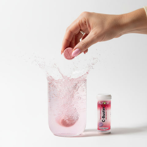 Hand dropping a pink tablet into a glass of water with a bottle labeled 'C-Buster' in the background.